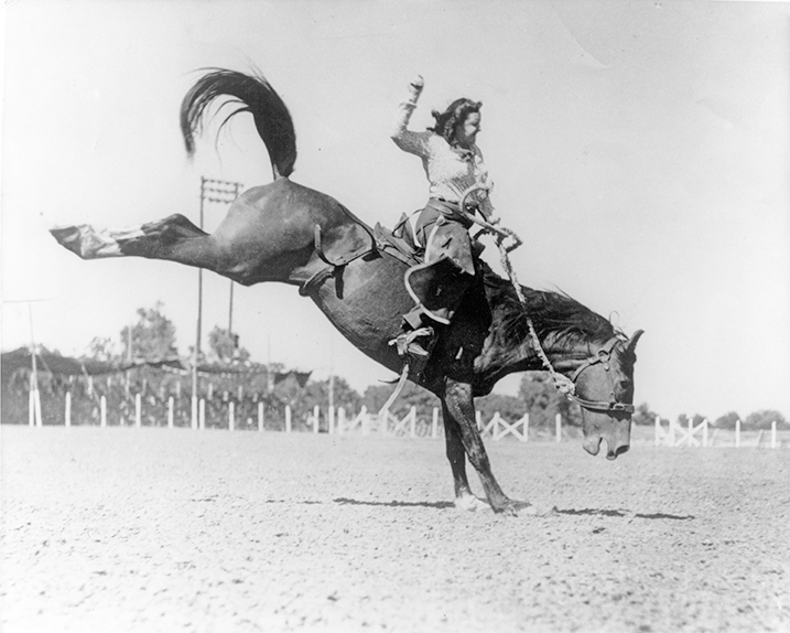 Cowboy on a bucking horse in an arena, rider raised arm while the horse rears, dusty ground and fencing in the background