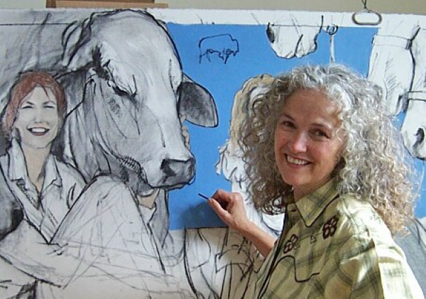 Artist with curly gray hair paints a cow mural on a blue wall, smiling at the camera.
