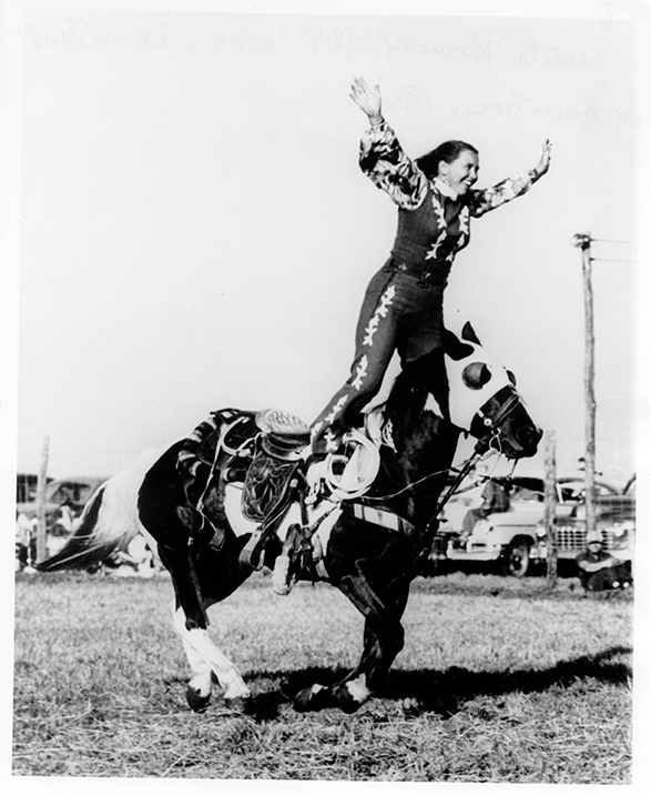 Rider balancing on a rearing horse with arms raised during a rodeo performance.