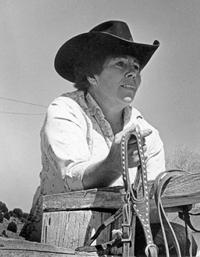 Woman in a cowboy hat leaning on a wooden fence, holding a lariat outdoors on a ranch.