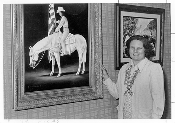 Woman in a light suit with a flower lei standing beside a framed painting of a cowboy on a white horse in a gallery.
