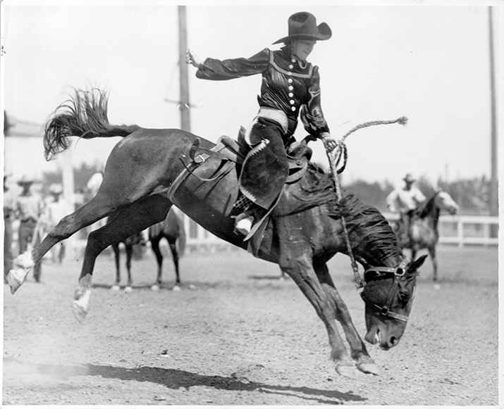 Cowgirl in a wide-brim hat riding a bucking bronc during a rodeo, horse mid-air and hooves kicked up on a dirt arena.
