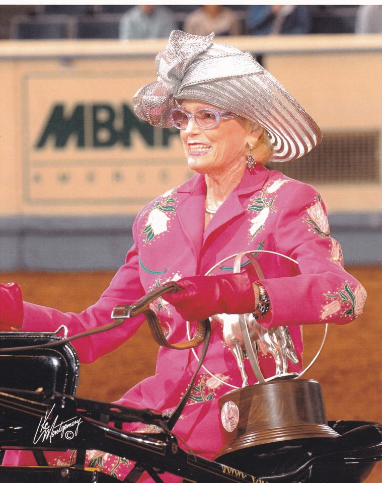 Smiling elderly woman in a glittering silver hat and pink embroidered suit rides a black motorcycle.