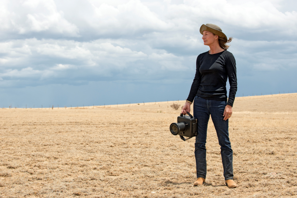 Woman standing in a dry, open field holding a large camera with a telephoto lens, wearing a beige hat and dark clothing, looking to the right.