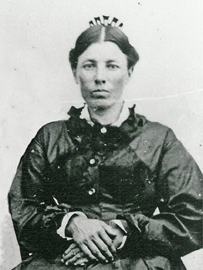 Portrait of a stern-looking woman in a dark 19th‑century dress, her hair up with a small comb, hands folded in her lap.