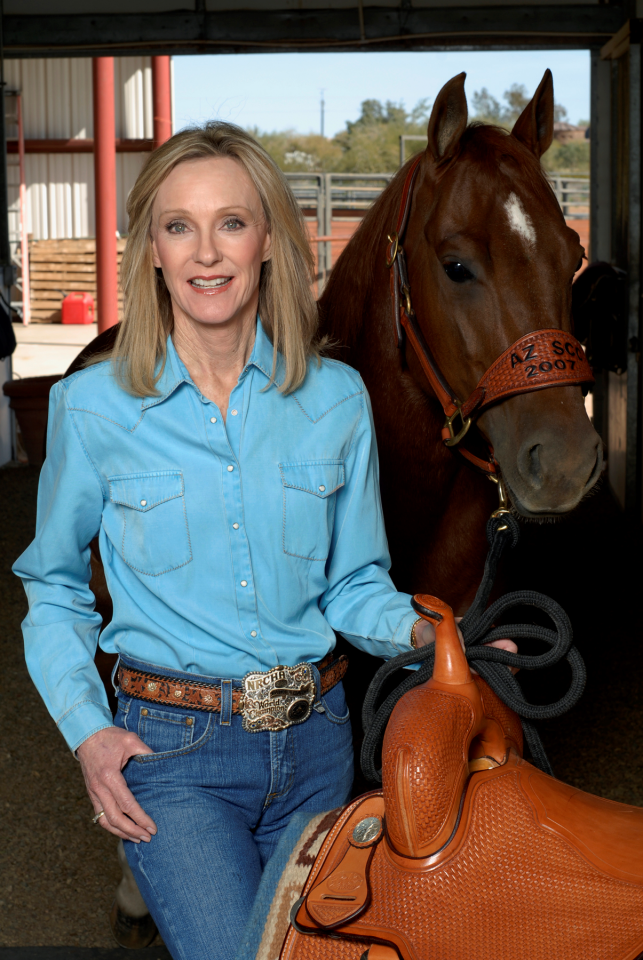 Smiling woman in a blue shirt standing beside a brown horse wearing a saddle in a stable.