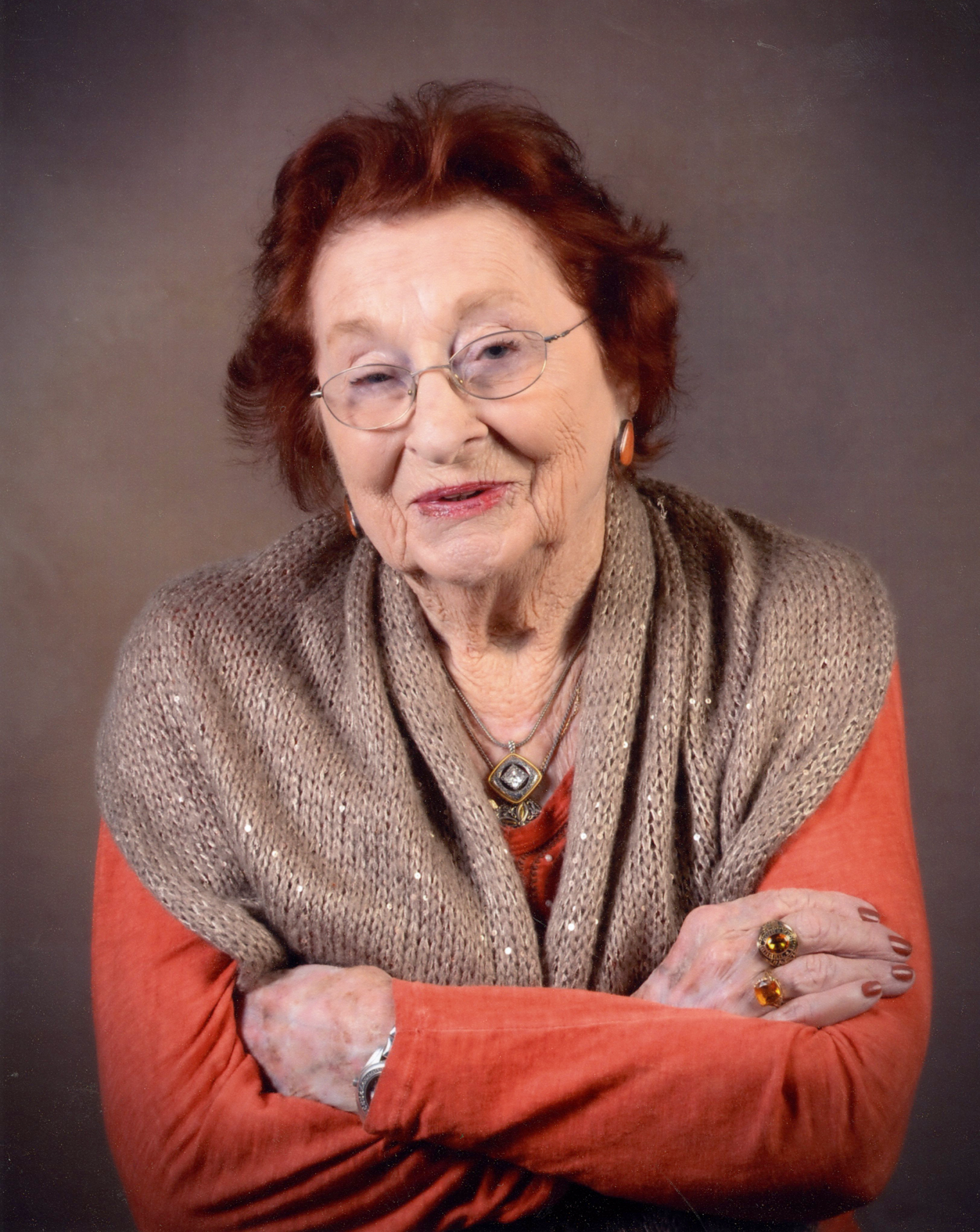 Portrait of an elderly woman with short reddish hair and glasses, wearing a brown knit scarf and orange sweater, arms crossed in a studio setting.