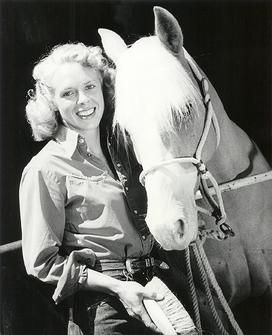 Smiling woman in a button-down shirt standing beside a white horse in a stable setting.