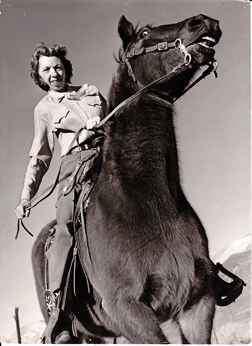 Smiling woman riding a bucking horse, holding the reins as the horse rears up outdoors in a sunny, mountainous setting.