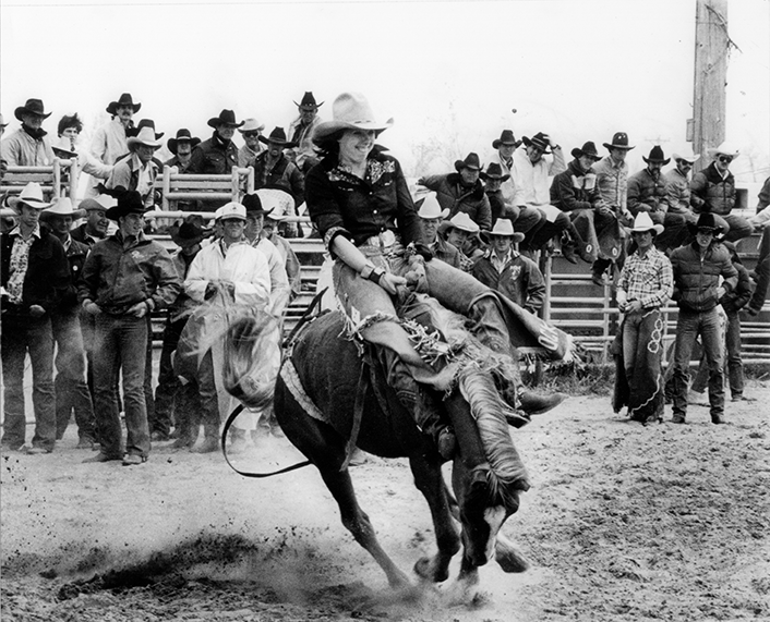 Female rodeo rider on a bucking horse, mid-air as dust rises, with spectators in hats watching from the stands.