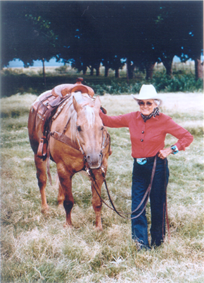 Older woman in a white hat and red jacket stands beside a saddled light-brown horse in a grassy field.