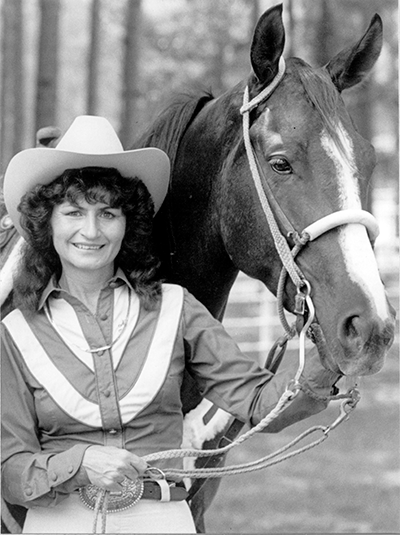 Woman in a cowboy hat smiles beside a horse, holding its reins in a paddock.