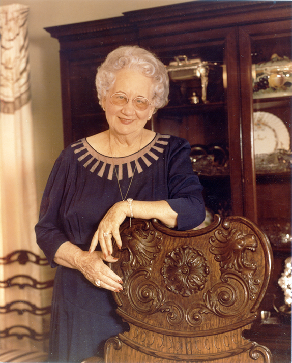 Smiling elderly woman with white curls and glasses, standing beside a carved wooden chair in a dining room.