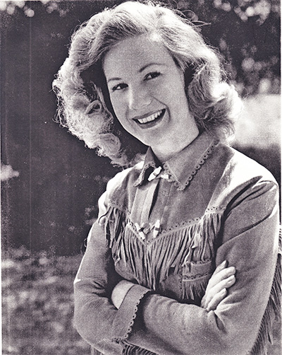 Smiling woman with wavy hair wearing a fringed western shirt, arms crossed, outdoors (black-and-white photo).