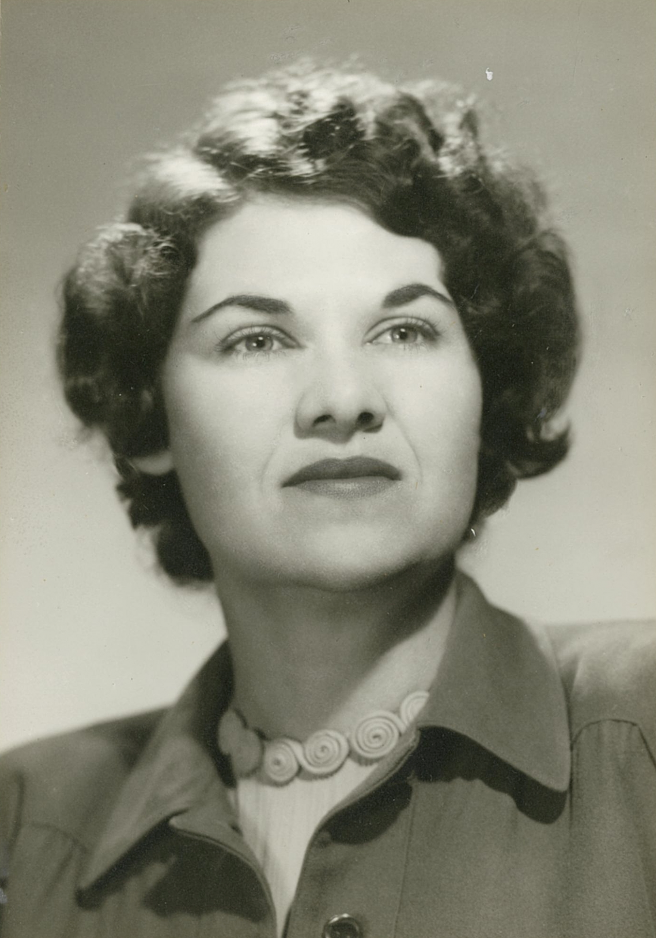 Close-up black-and-white portrait of a woman looking upward, with short, wavy hair and a beaded necklace.