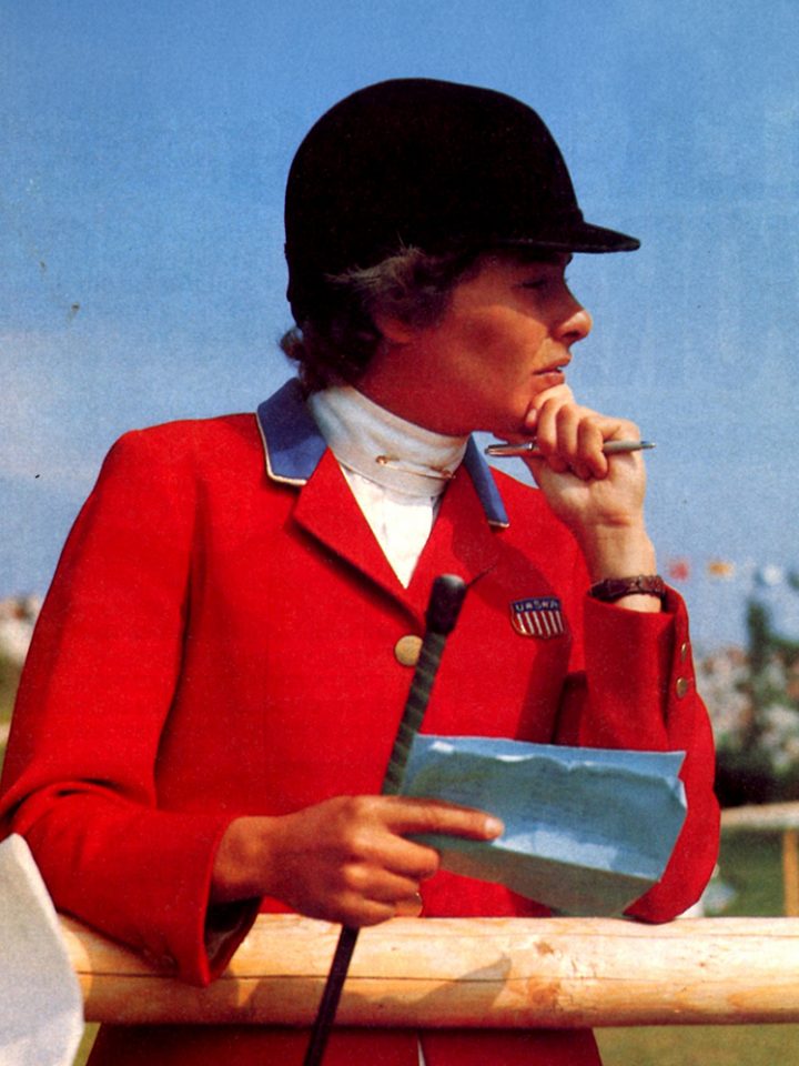 Person in a red blazer with an American patch, black cap, resting chin on hand while holding a pen and a notebook.