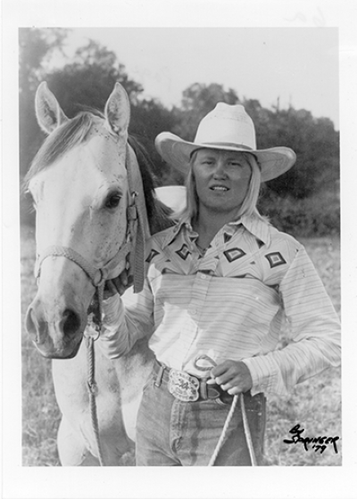 A woman in a white cowboy hat and patterned shirt stands beside a white horse outdoors, holding the reins.