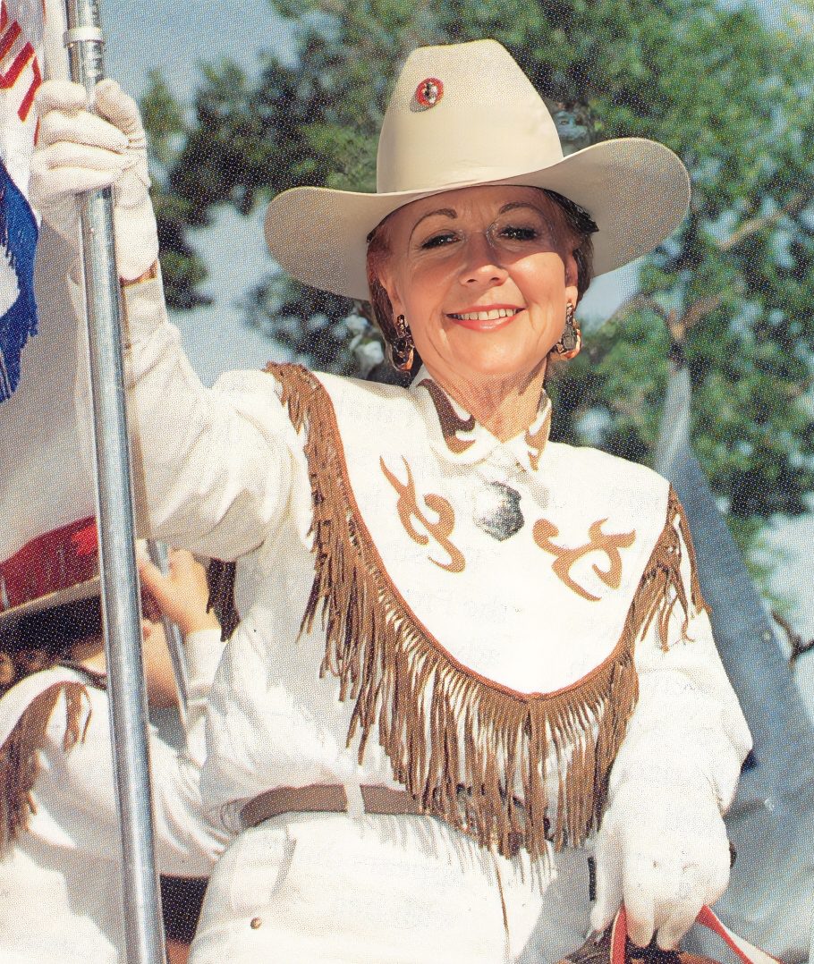 Smiling woman in a white fringed western shirt and wide-brimmed hat at a parade, holding a flagpole beside her.