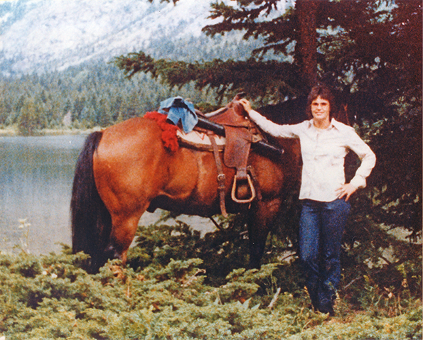 Person standing beside a saddled brown horse near a lake with trees and mountains in the background.