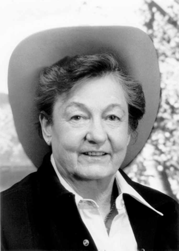 Older woman wearing a wide-brimmed hat, smiling in a blazer and white shirt (black-and-white portrait).
