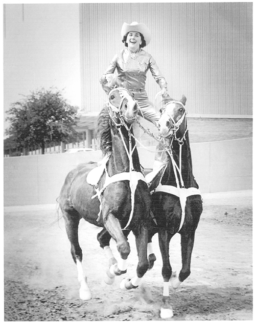 Rider in a cowboy hat balances on two competing horses side by side, performing a stunt in an arena while smiling.
