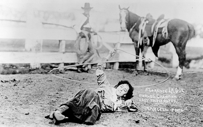 Black-and-white photo of Florence LaDue posing on the dirt with a lasso, two saddled horses in the background, suggesting a rodeo or trick-roping scene.