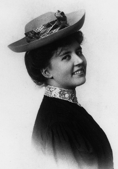 Portrait of a smiling woman wearing a vintage wide-brim hat with a plaid ribbon and lace collar, in a black-and-white studio photo.