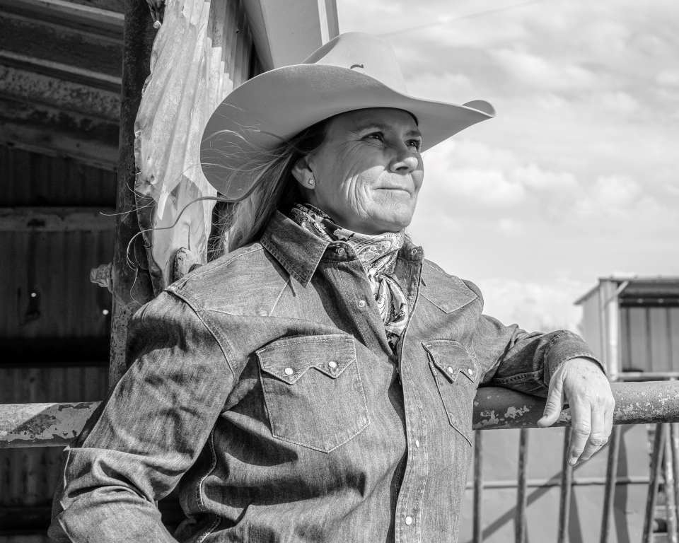 Outdoor black-and-white portrait of a person wearing a wide-brimmed cowboy hat, denim shirt, and bandana, leaning on a railing.