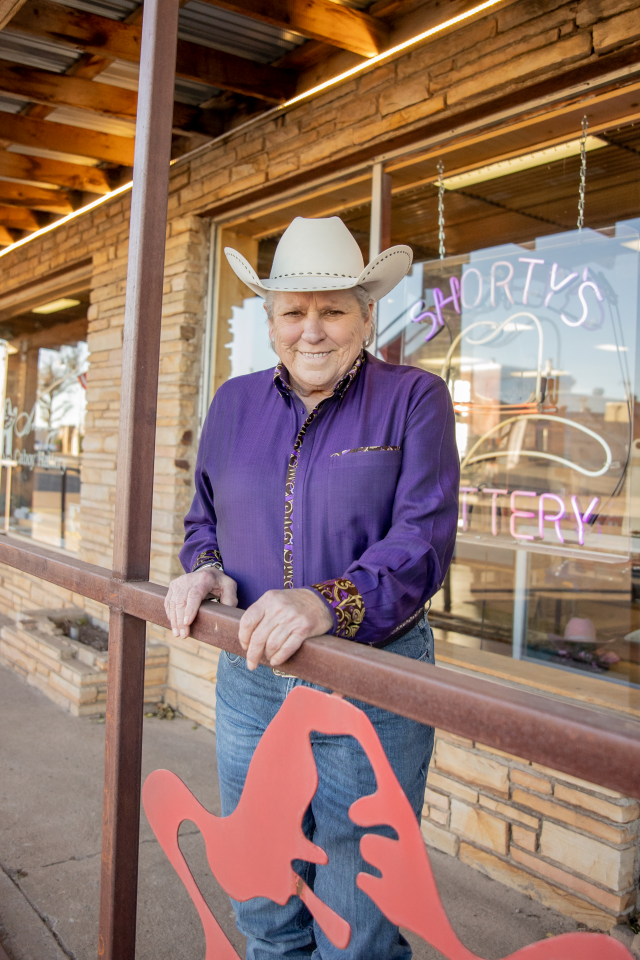Smiling older man in a white cowboy hat and purple shirt leaning on a railing outside a brick storefront with neon signage.