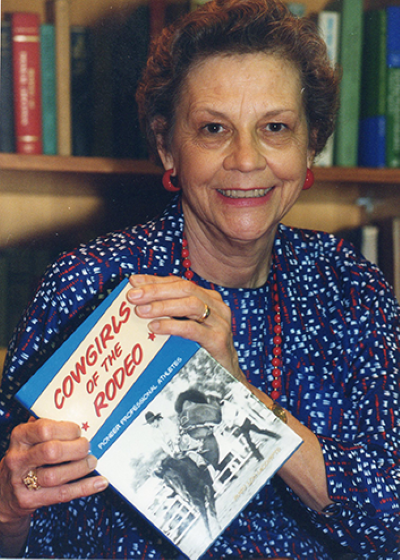 Older woman with short curly hair, wearing a blue patterned jacket and red earrings, smiles while holding a book titled 'Cowgirls of the Rodeo' in a library setting.