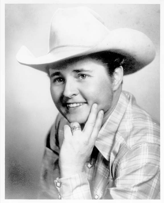 Black-and-white portrait of a person in a cowboy hat and plaid shirt, smiling with a hand resting at the chin.