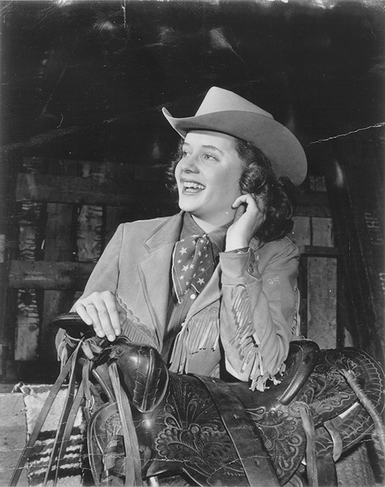 Smiling woman in a cowboy hat and fringe jacket sits on a saddle, resting her hand on her cheek in a rustic setting.