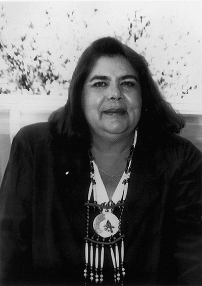 Portrait of a middle-aged woman with dark hair wearing a blazer and a beaded necklace, seated indoors.