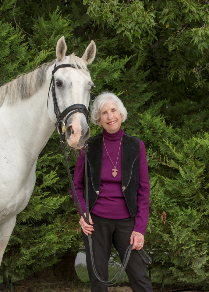 Elderly woman in a purple top and black vest stands beside a white horse, smiling, with evergreen shrubs behind.