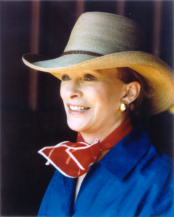 Smiling woman wearing a wide-brim straw hat, red scarf, and blue jacket with gold earrings.