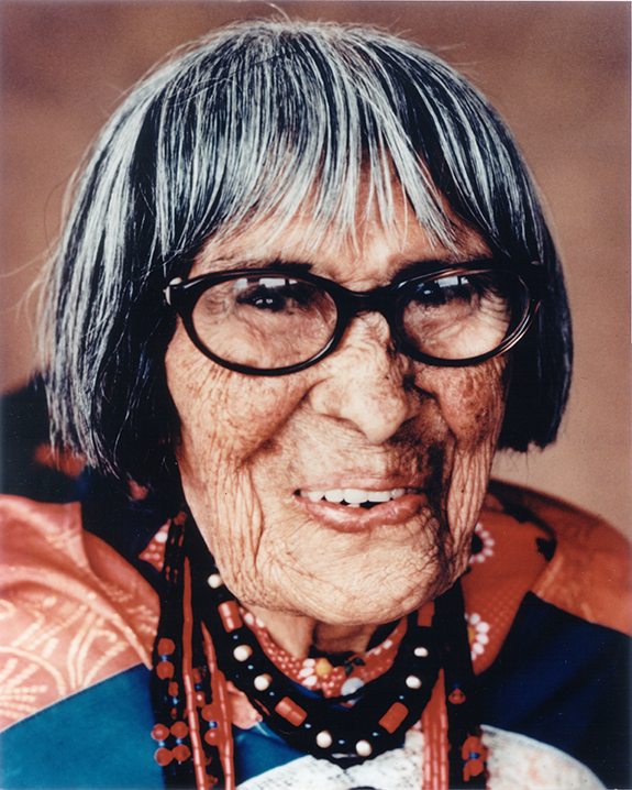 Portrait of an elderly Indigenous woman with black-framed glasses, gray-black hair, and beaded necklace, smiling gently in colorful clothing.