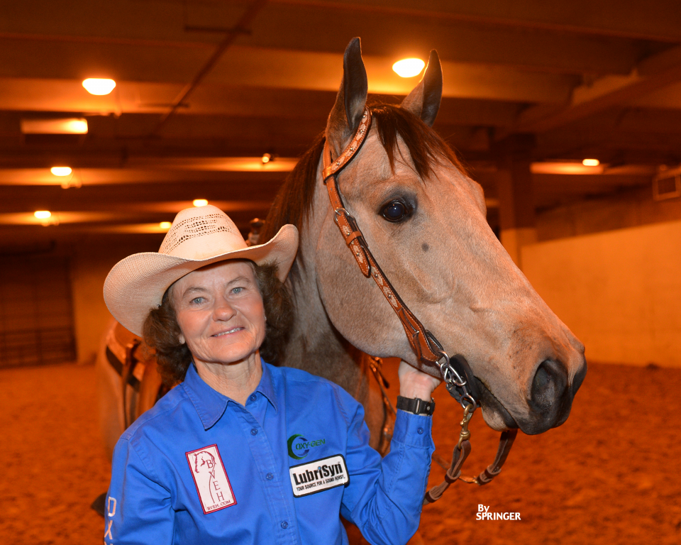 A smiling woman in a cowboy hat and blue shirt stands beside a brown horse in an indoor arena, her hand gently holding the horses bridle.