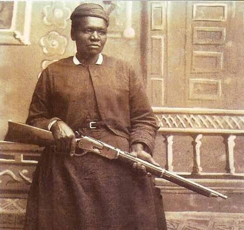 Sepia-toned photo of a Black woman in a long dress, standing on a porch and holding a rifle across her arms.
