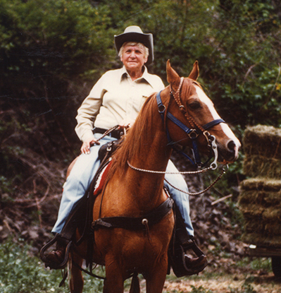 Older man wearing a tan shirt and hat riding a brown horse along a dirt path with green vegetation and straw bales in the background.