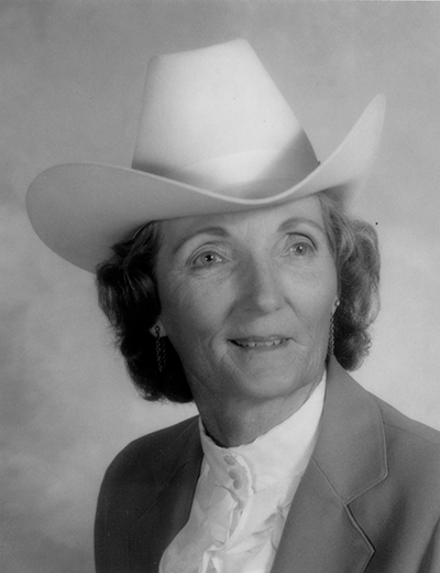 Portrait of an older woman in a white cowboy hat, blazer, and ruffled blouse, looking slightly to the right in a studio portrait.