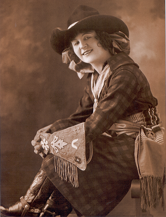 Vintage sepia portrait of a smiling woman in a wide-brimmed hat and Western outfit, seated with decorative fringed shawl and boots.
