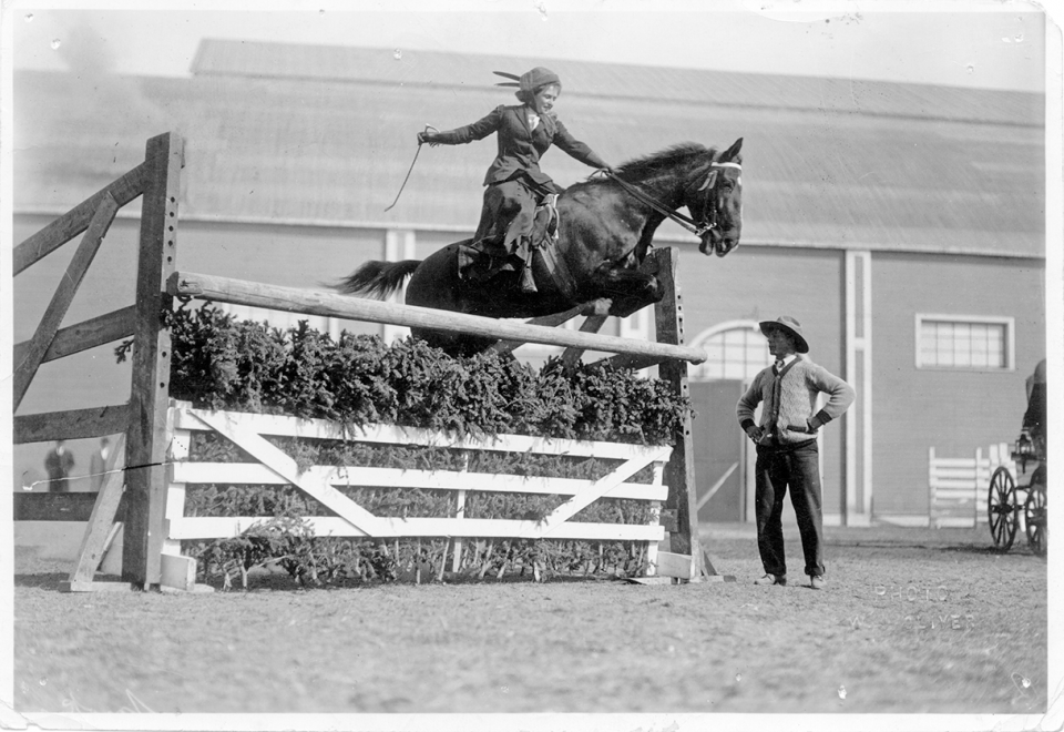 Horse rider clears a jump at a show jumping event, while a man in a hat watches nearby; wooden fence and shrubs form the obstacle.