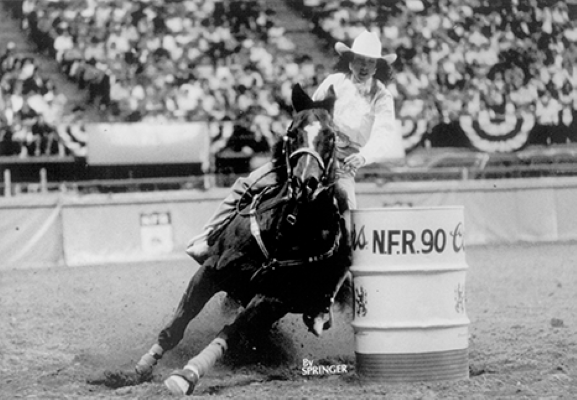 Black-and-white photo of a cowboy on a horse turning around a barrel at a rodeo arena, with spectators in the stands behind.