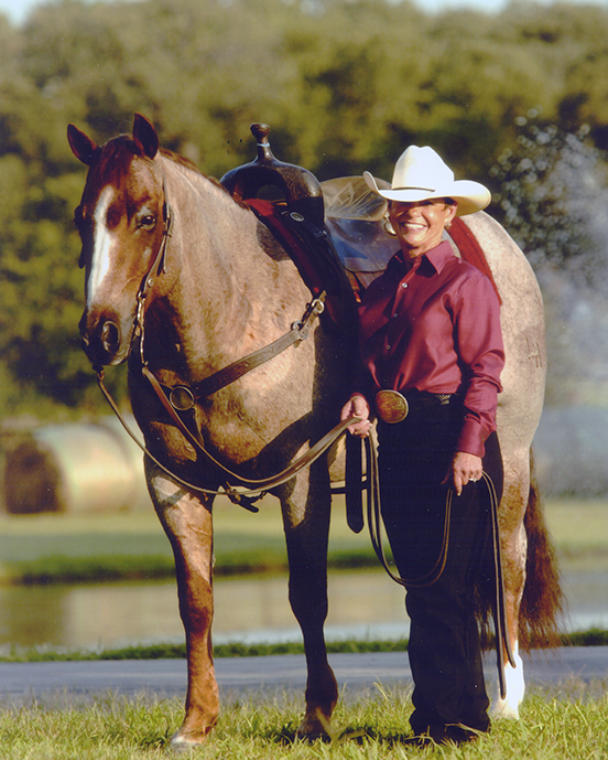 Smiling cowboy in a white hat stands beside a saddled brown horse outdoors on a grassy field.
