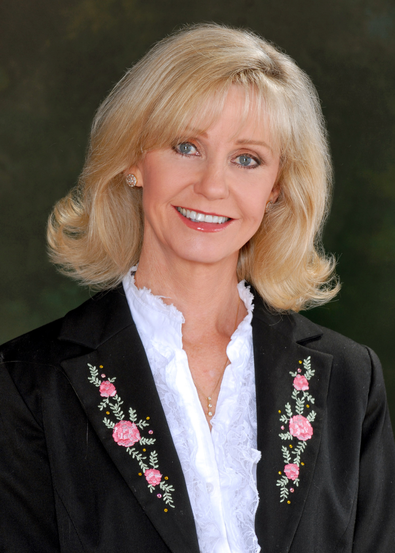 Portrait of a smiling blonde woman wearing a black embroidered blazer and white blouse outdoors