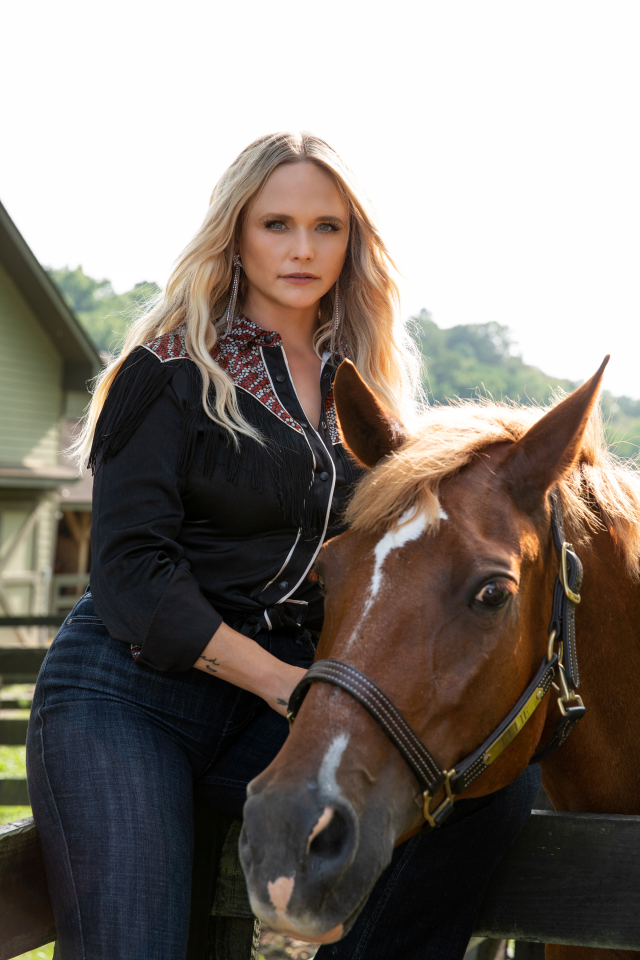Woman with long blonde hair riding a horse, wearing a black fringed shirt and jeans outdoors at a ranch/stables.