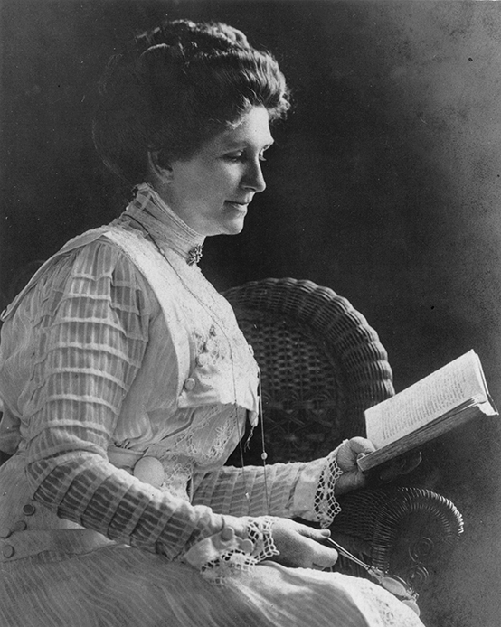 Woman in a lace dress sits in a wicker chair, reading a book from her lap.