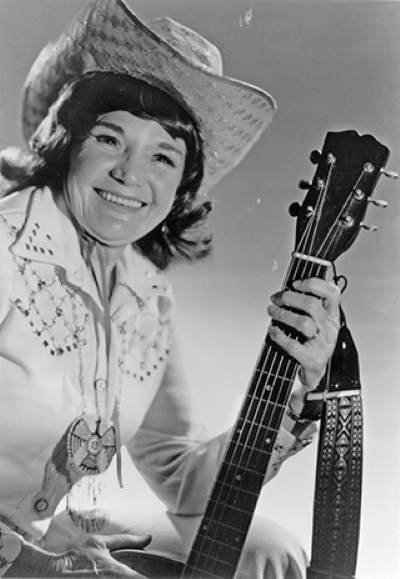 Black-and-white photo of a smiling woman in a cowboy hat holding an acoustic guitar close to her body.