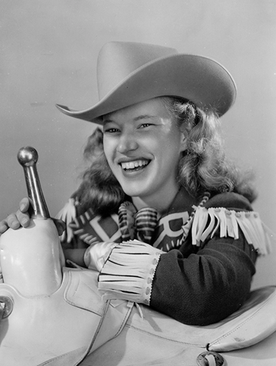 Young woman wearing a cowboy hat and fringed jacket smiles while holding the reins of a white mechanical carousel horse.