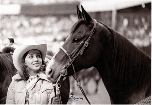 Woman wearing a white cowboy hat and light jacket stands beside a dark horse, looking at it while holding its reins at a rodeo or show.